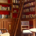 comfortable chairs, a table, surrounded by shelves of books with a book ladder. A window floods the room with light.